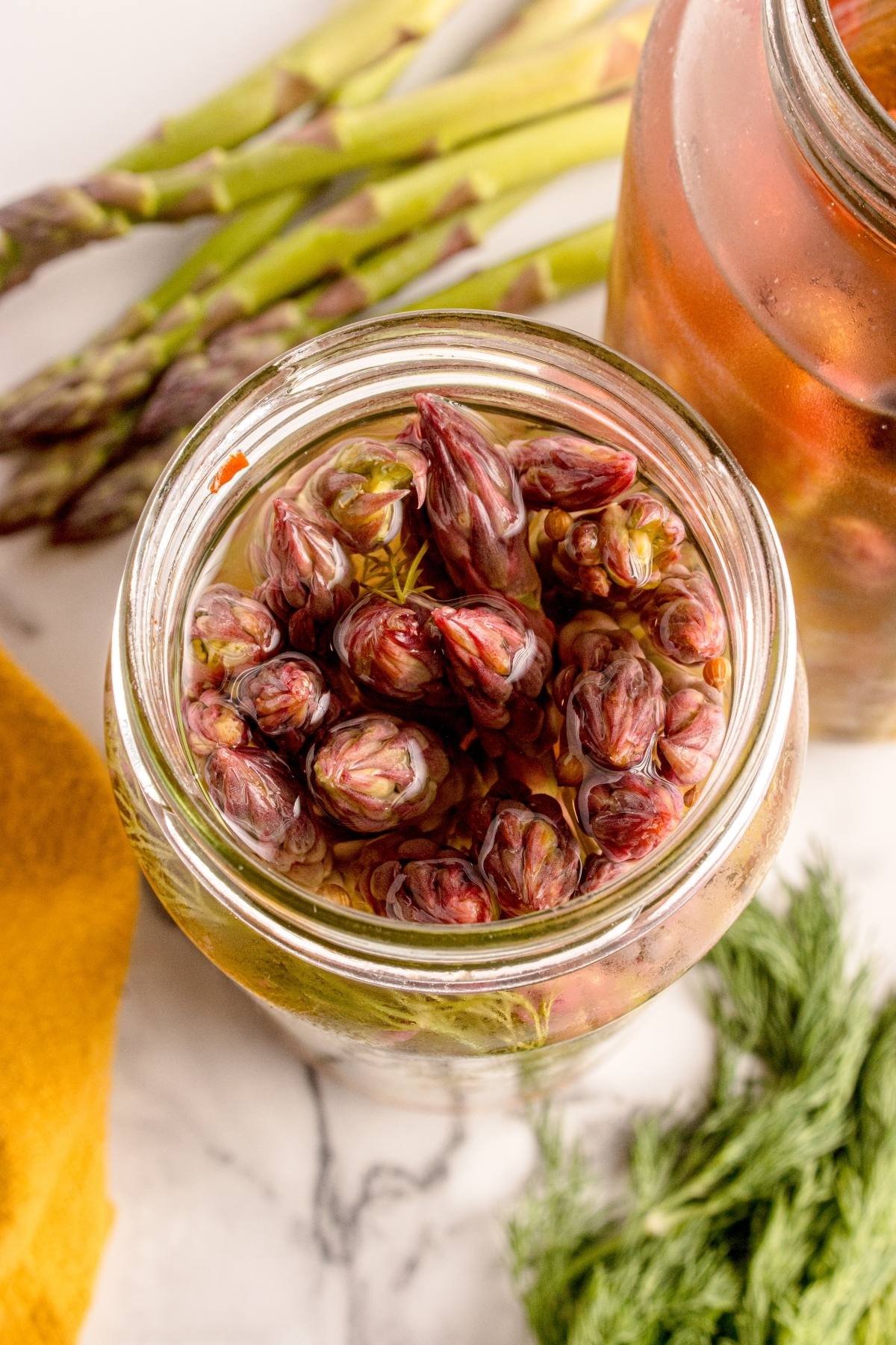 A jar filled with pickled asparagus tips, surrounded by fresh asparagus stalks and dill on a marble surface.