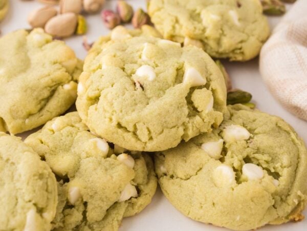 Pistachio Pudding Cookies with white chocolate chips, surrounded by pistachios on a white surface.