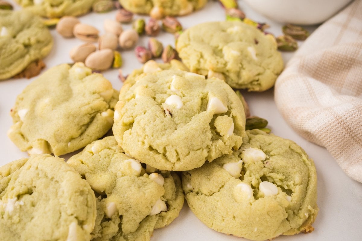 Pistachio Pudding Cookies with white chocolate chips sit on a countertop, surrounded by shelled pistachios.