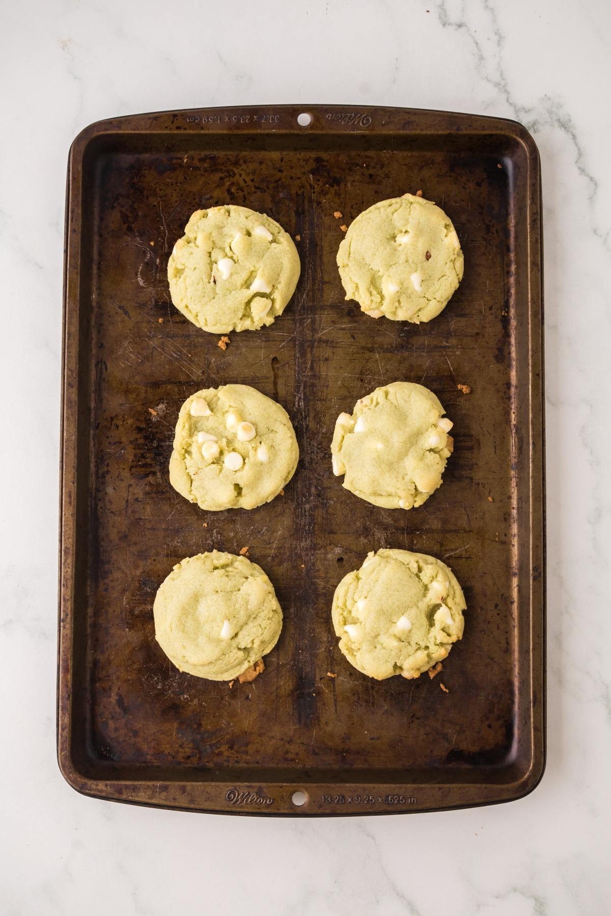 Six baked cookies with white chocolate chips on a dark baking sheet, placed on a white marble surface.