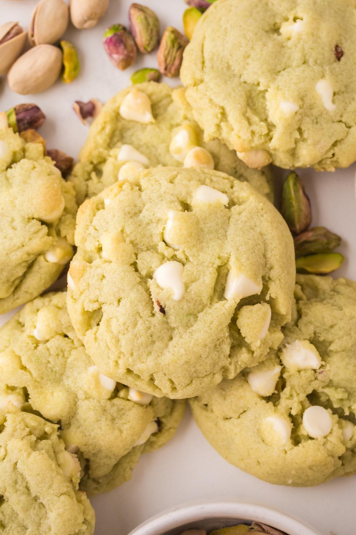 Pistachio cookies with white chocolate chips, scattered with whole pistachios on a white surface.