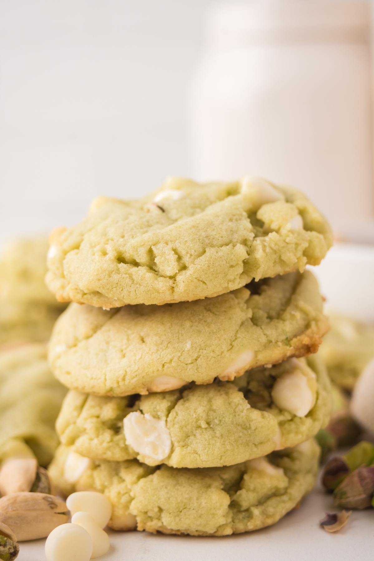 A stack of green cookies with white chocolate chips, surrounded by pistachios and a jar in the background.
