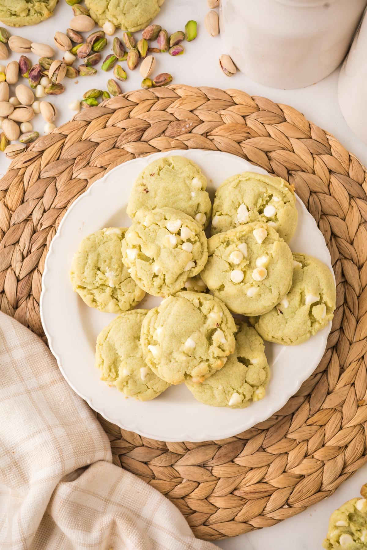 A plate of green pistachio cookies with white chocolate chips on a woven mat, surrounded by pistachios.