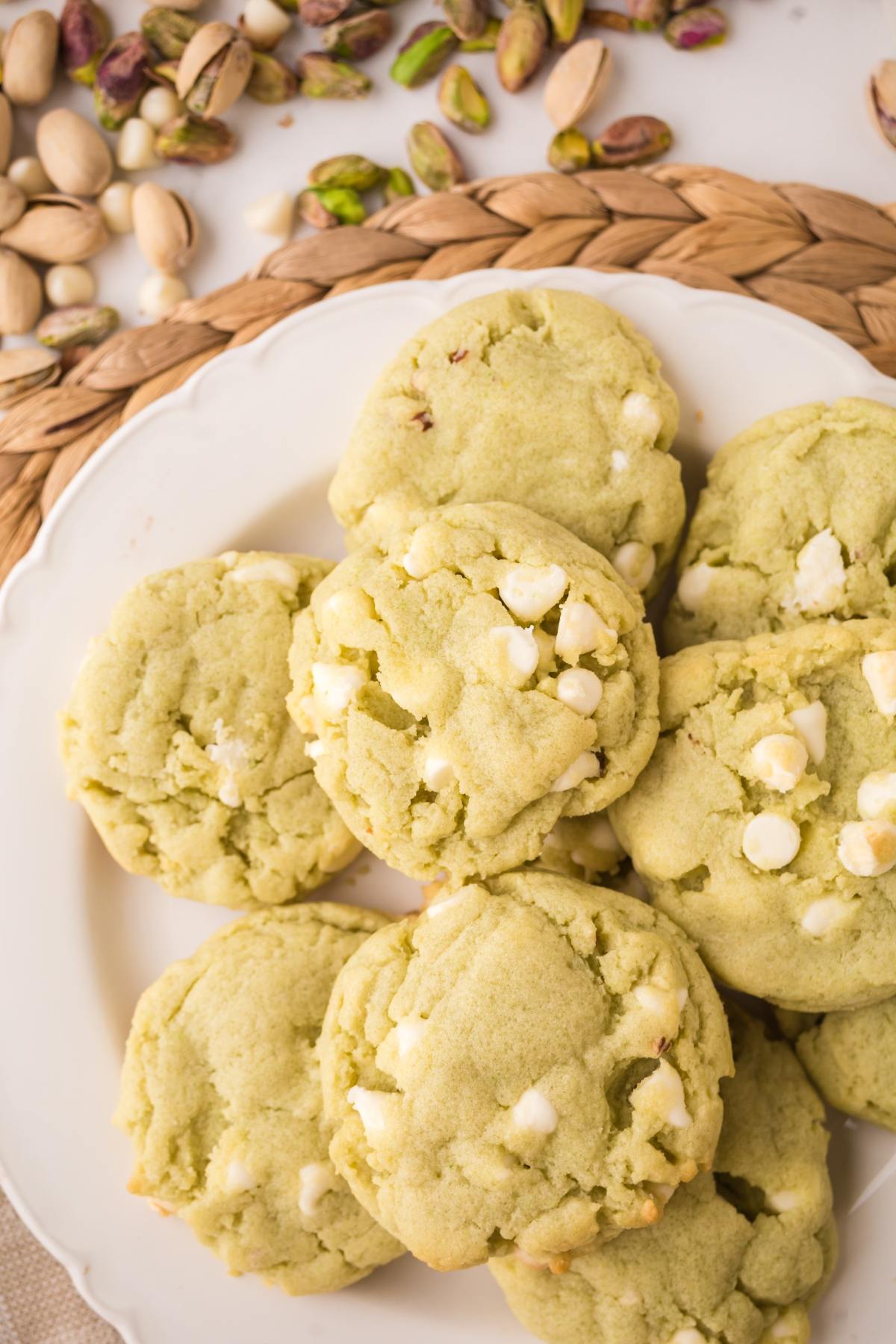 A plate of green pistachio cookies with white chocolate chips, surrounded by shelled and unshelled pistachios.