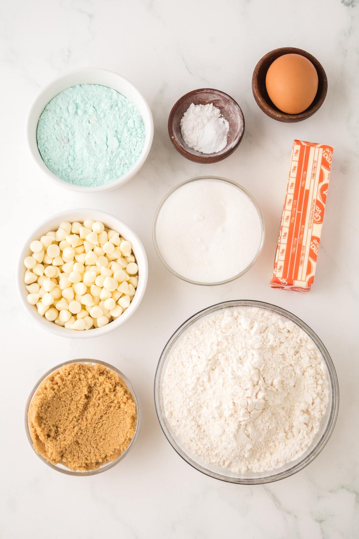 Baking ingredients in bowls: flour, sugar, brown sugar, white chips, blue powder, baking soda, butter, and an egg.