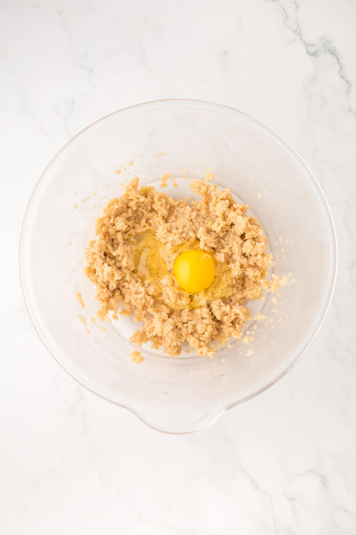 A clear bowl with creamed cookie dough and a raw egg in the center on a white marble surface.