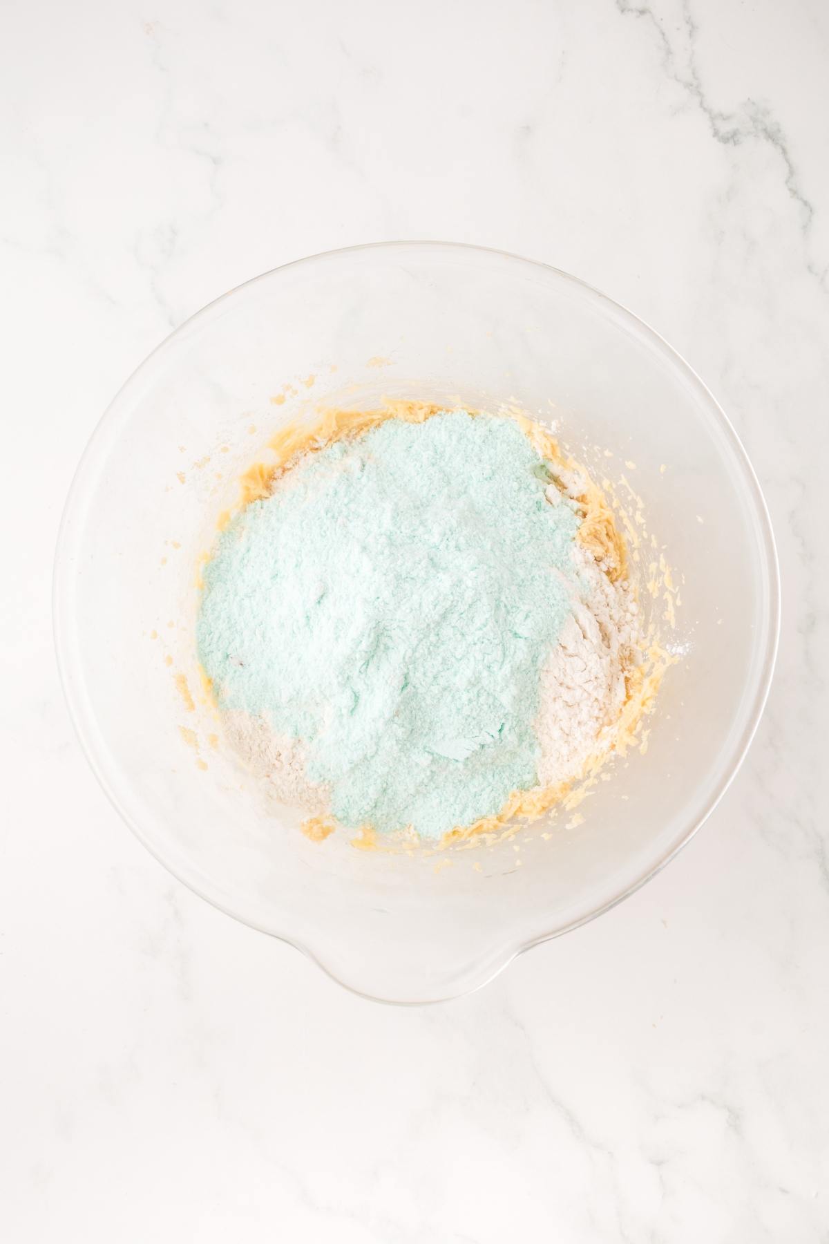 A clear bowl with baking ingredients, including a pile of blue powder, on a white marble surface.