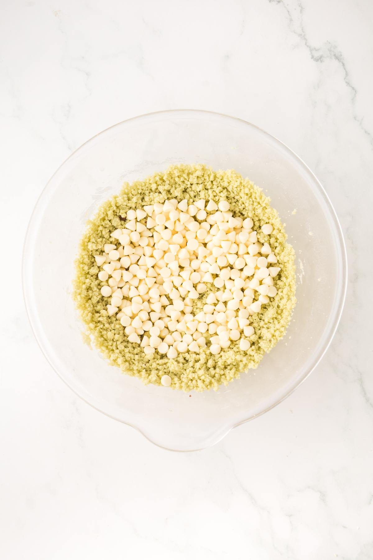A glass bowl with green cookie dough and white chocolate chips on a white marble surface.