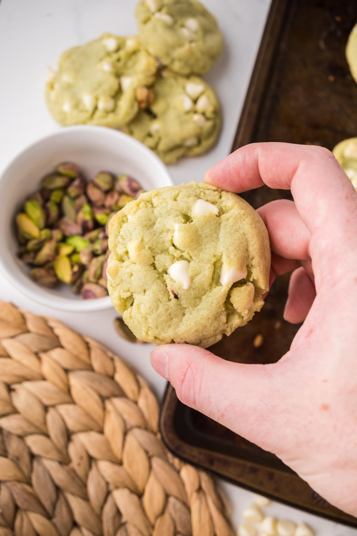 A hand holds a white chocolate pistachio cookie above a bowl of pistachios and a baking sheet.