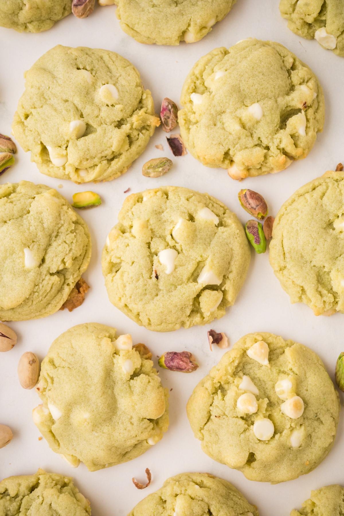 Green pistachio cookies with white chocolate chips, surrounded by scattered shelled pistachios on a white surface.