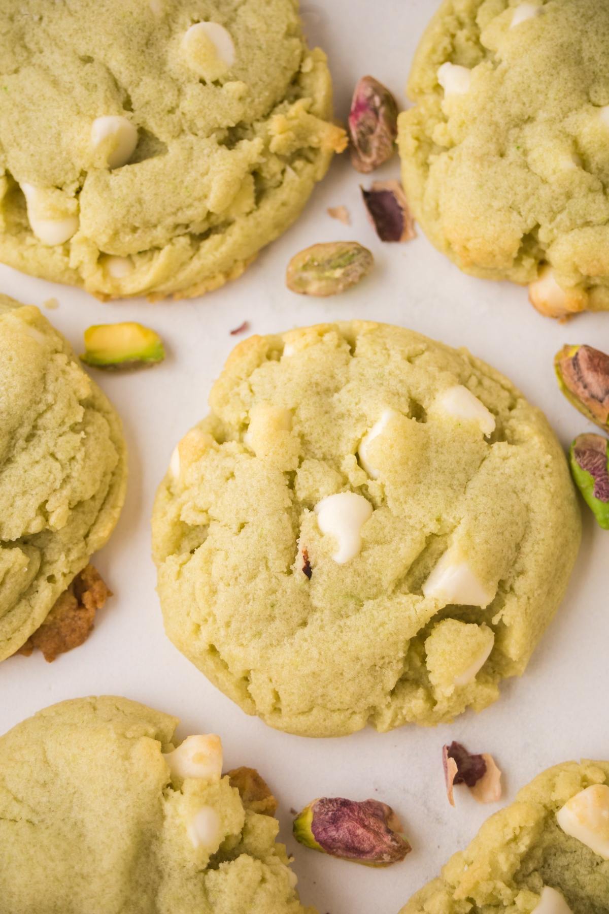 Soft green cookies with white chocolate chips and scattered pistachio nuts on a white surface.