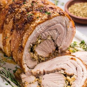 Porchetta roast with crispy skin and savory stuffing, sliced on a platter, garnished with fresh herbs and served alongside a bowl of seasoning.