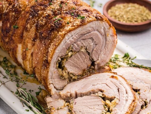 Porchetta roast with crispy skin and savory stuffing, sliced on a platter, garnished with fresh herbs and served alongside a bowl of seasoning.