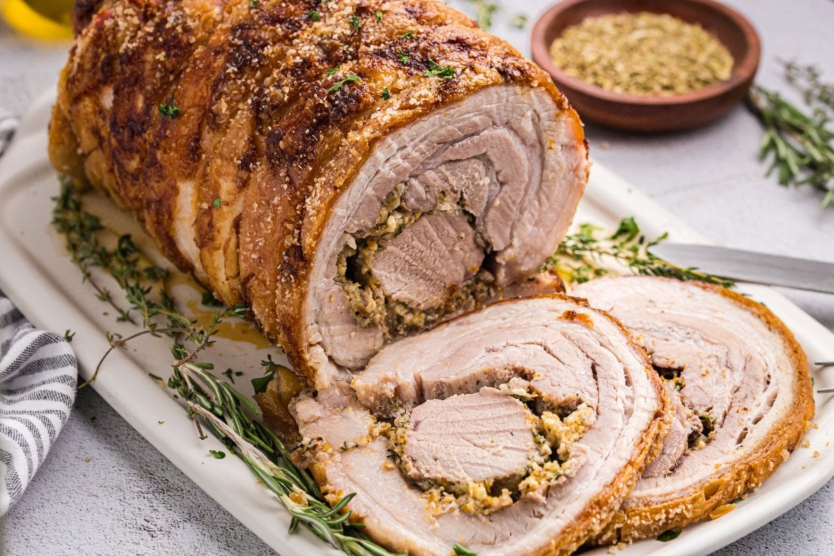 Sliced porchetta roast on a white platter, garnished with fresh herbs, with seasoning in the background.