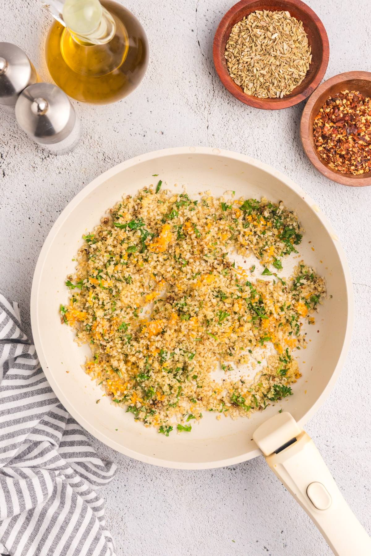 A white skillet with toasted bread crumbs, herbs, and orange zest, next to spices and olive oil on a gray surface.