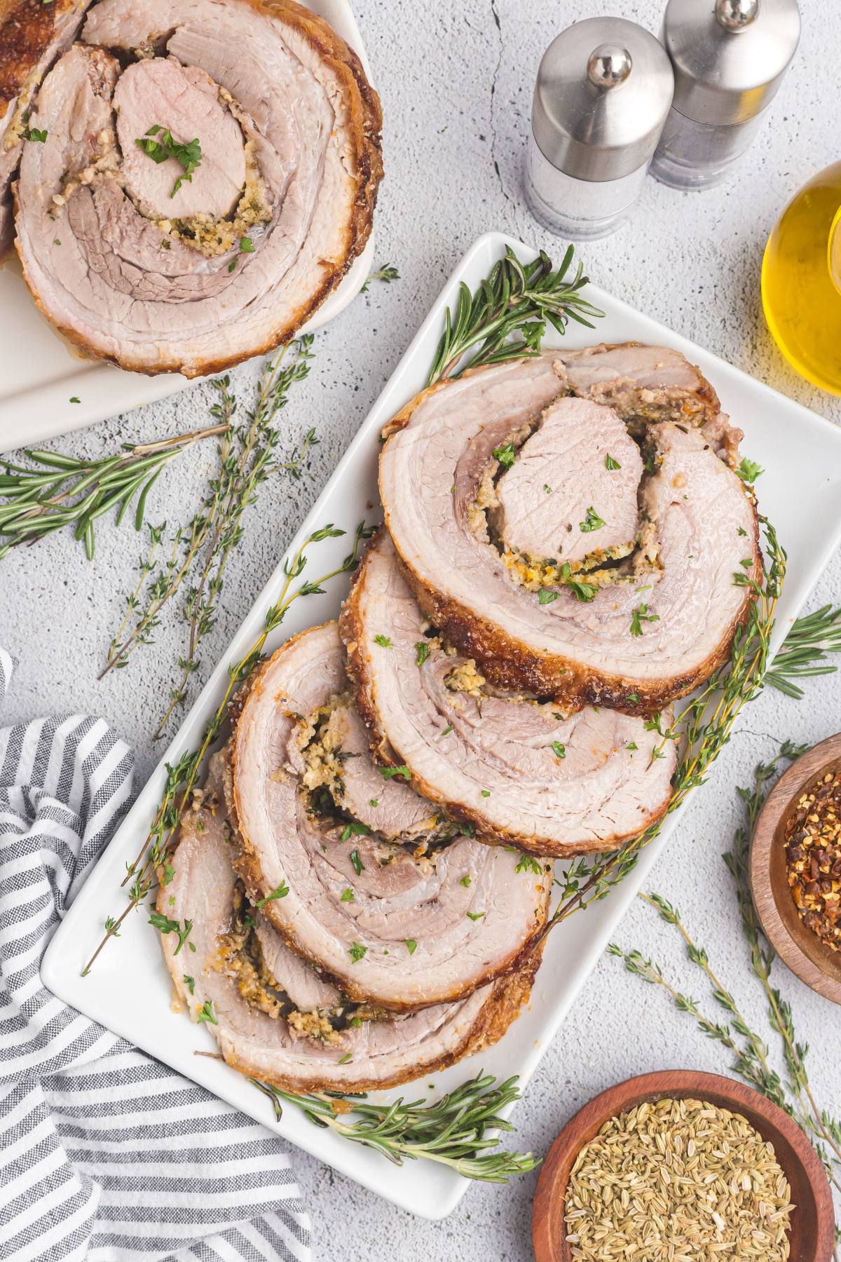 Sliced roast pork garnished with herbs on a white plate, surrounded by spices and seasonings.