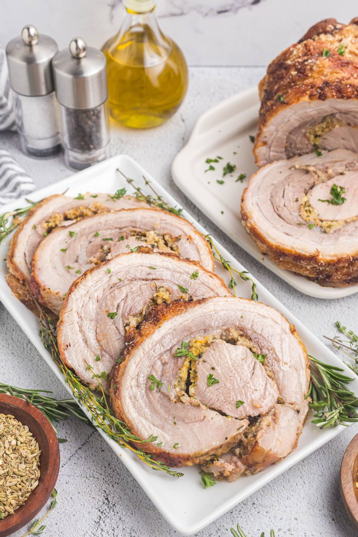 Three slices of stuffed roasted pork on a white plate, garnished with herbs, with whole roast and condiments nearby.