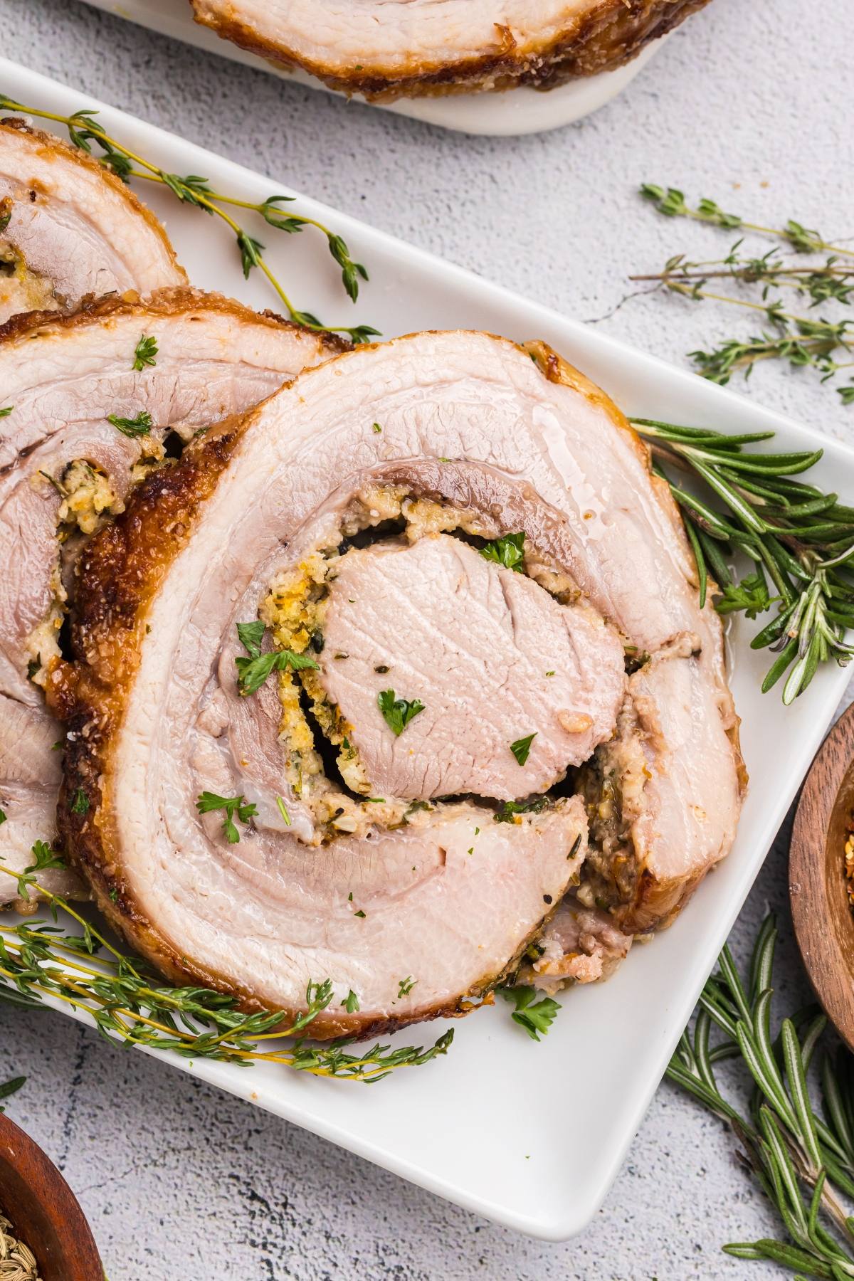 Sliced stuffed pork roast garnished with fresh rosemary and parsley on a white plate.