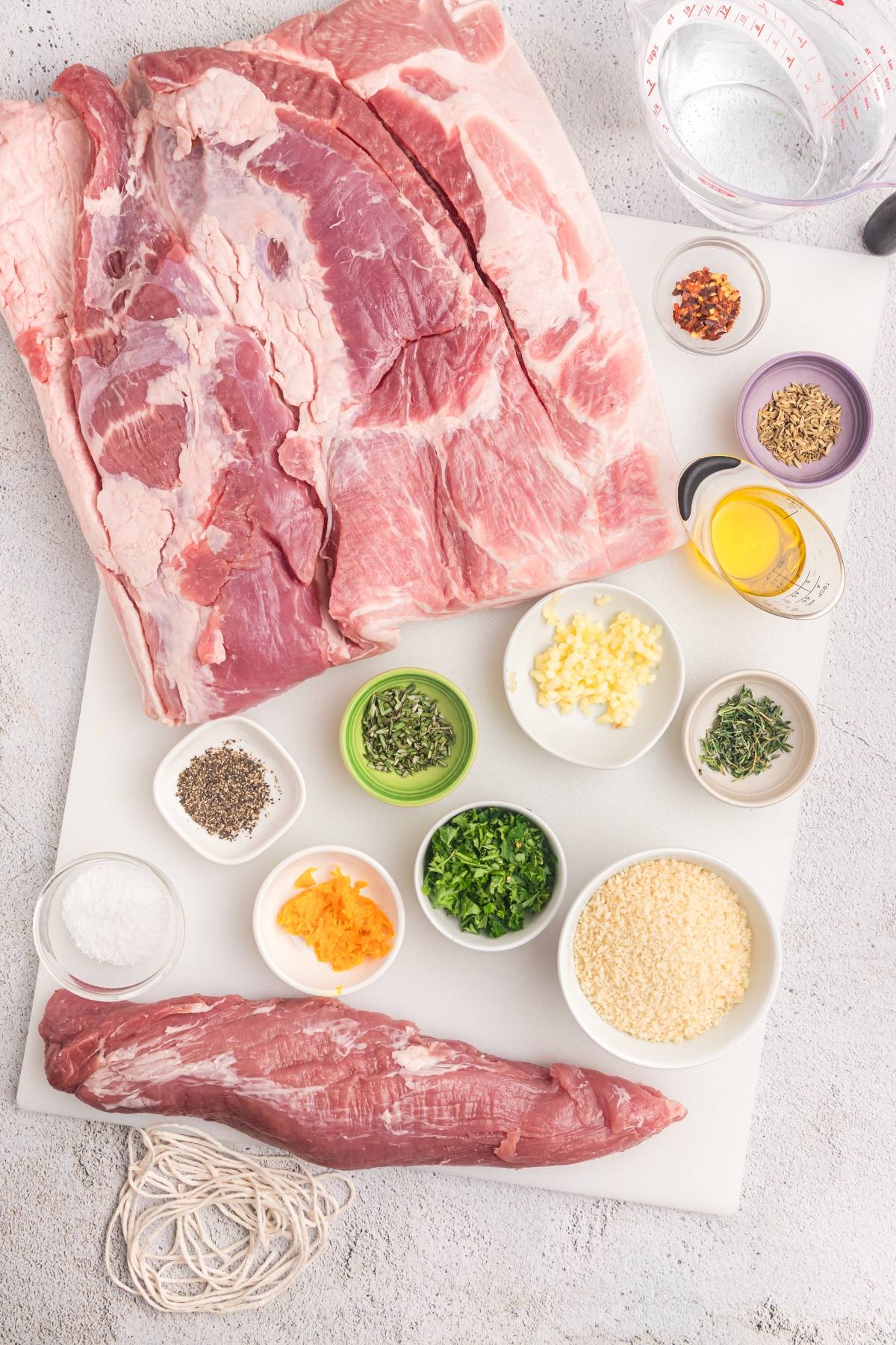 Raw pork cuts, herbs, spices, oil, and other ingredients arranged in bowls on a white cutting board.
