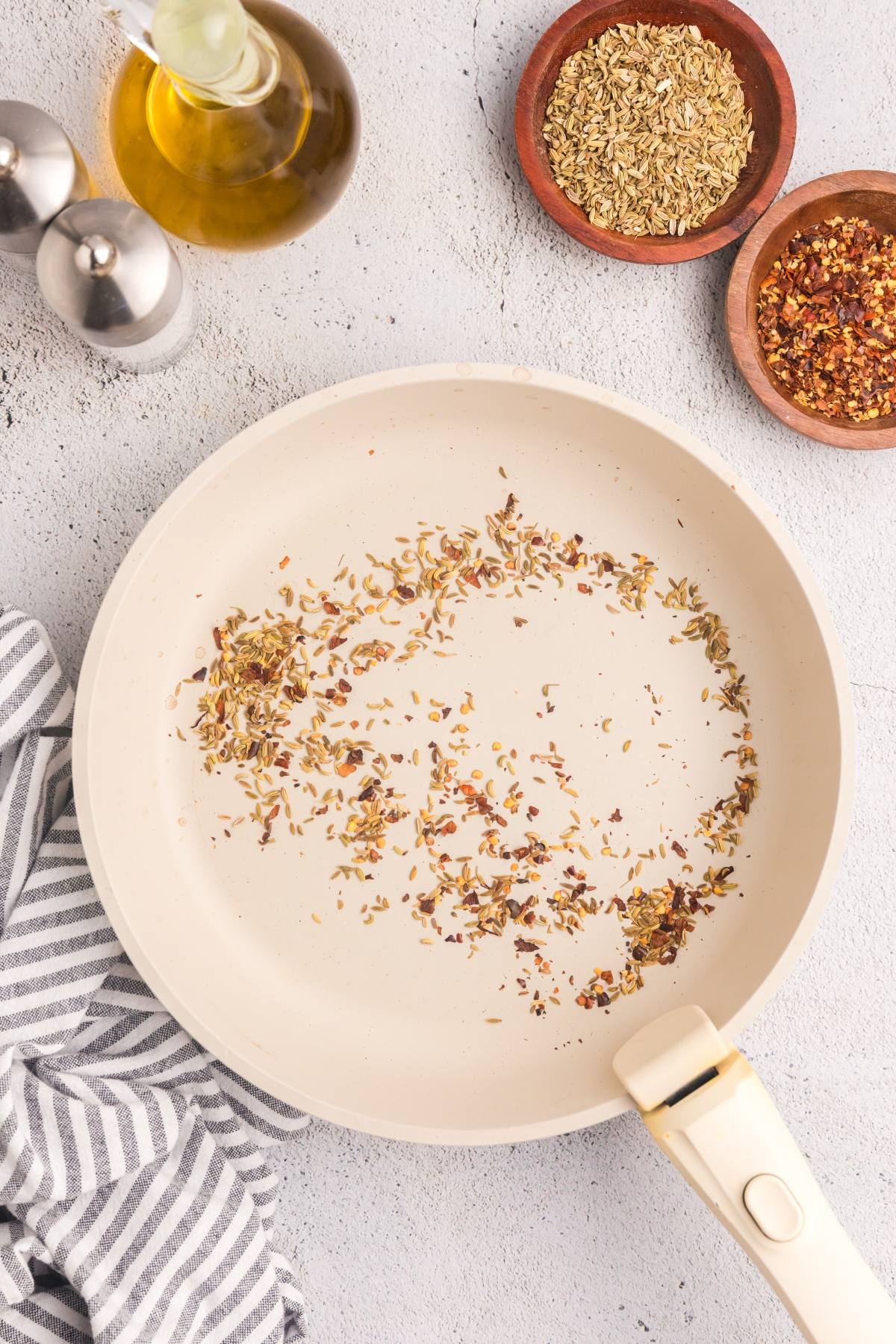 A frying pan with scattered dried herbs, next to bowls of spices, olive oil, and a striped towel.