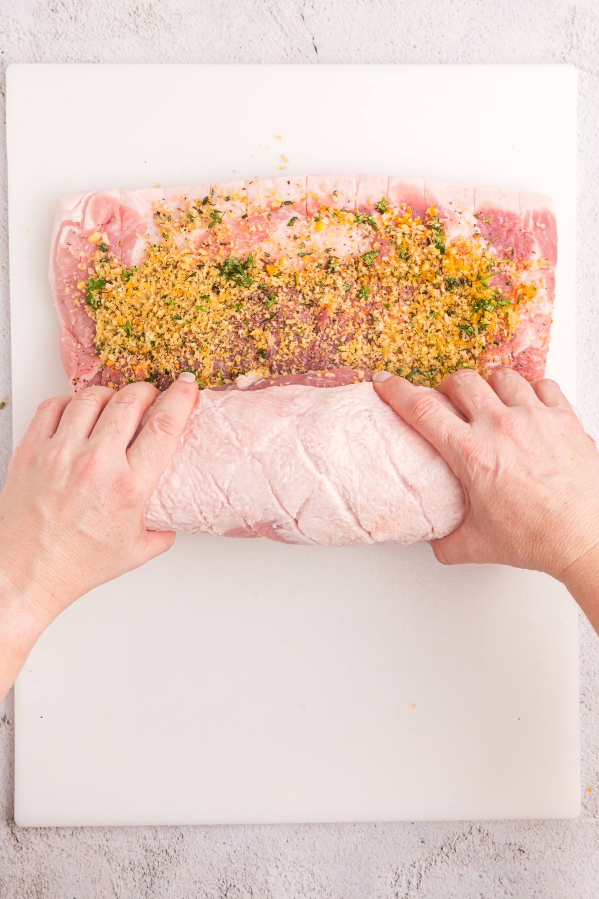 Hands rolling pork loin with herb and breadcrumb stuffing on a white cutting board.