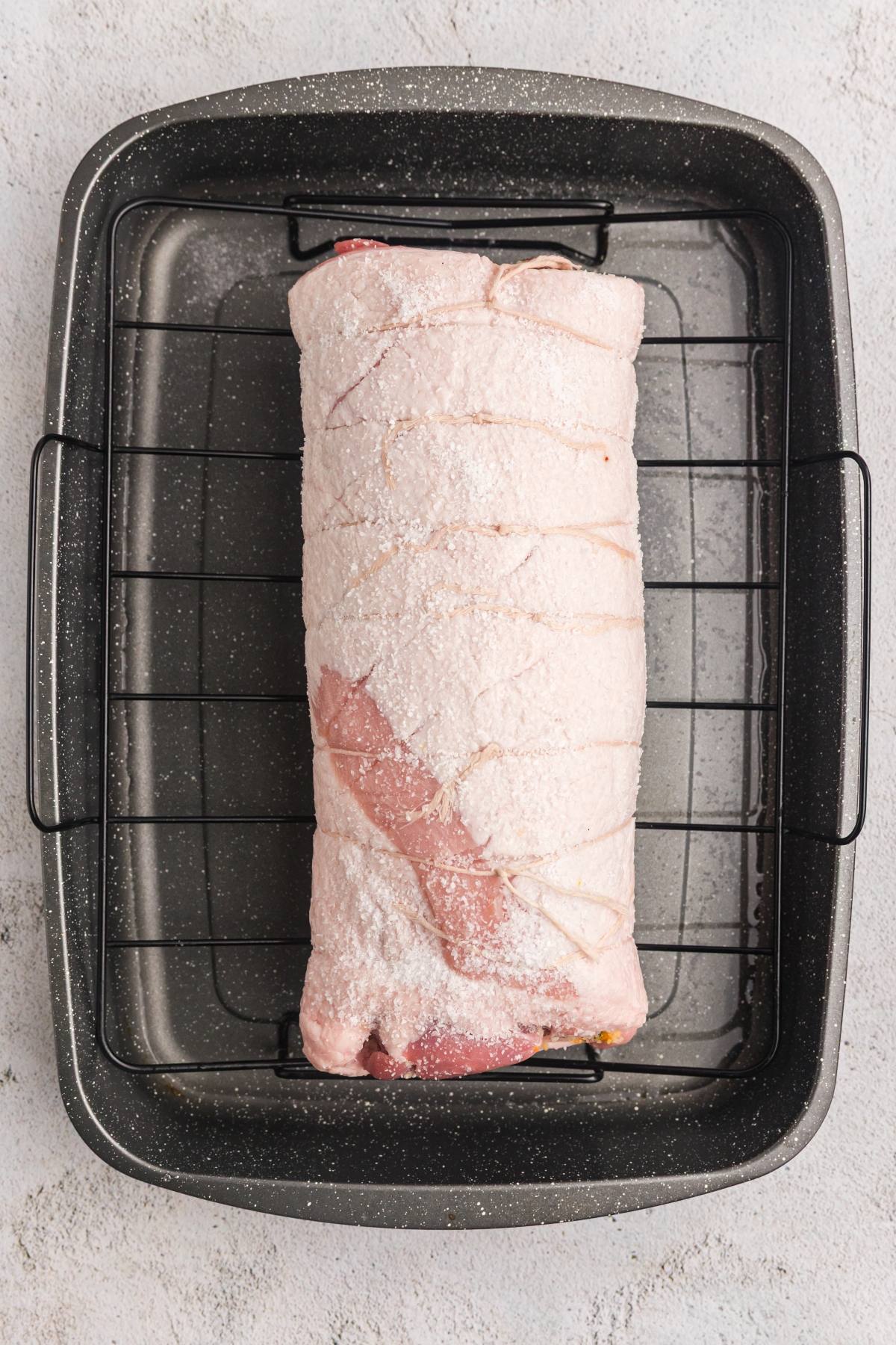 Uncooked pork roast covered in salt, tied with string, placed in a black roasting pan with a wire rack.
