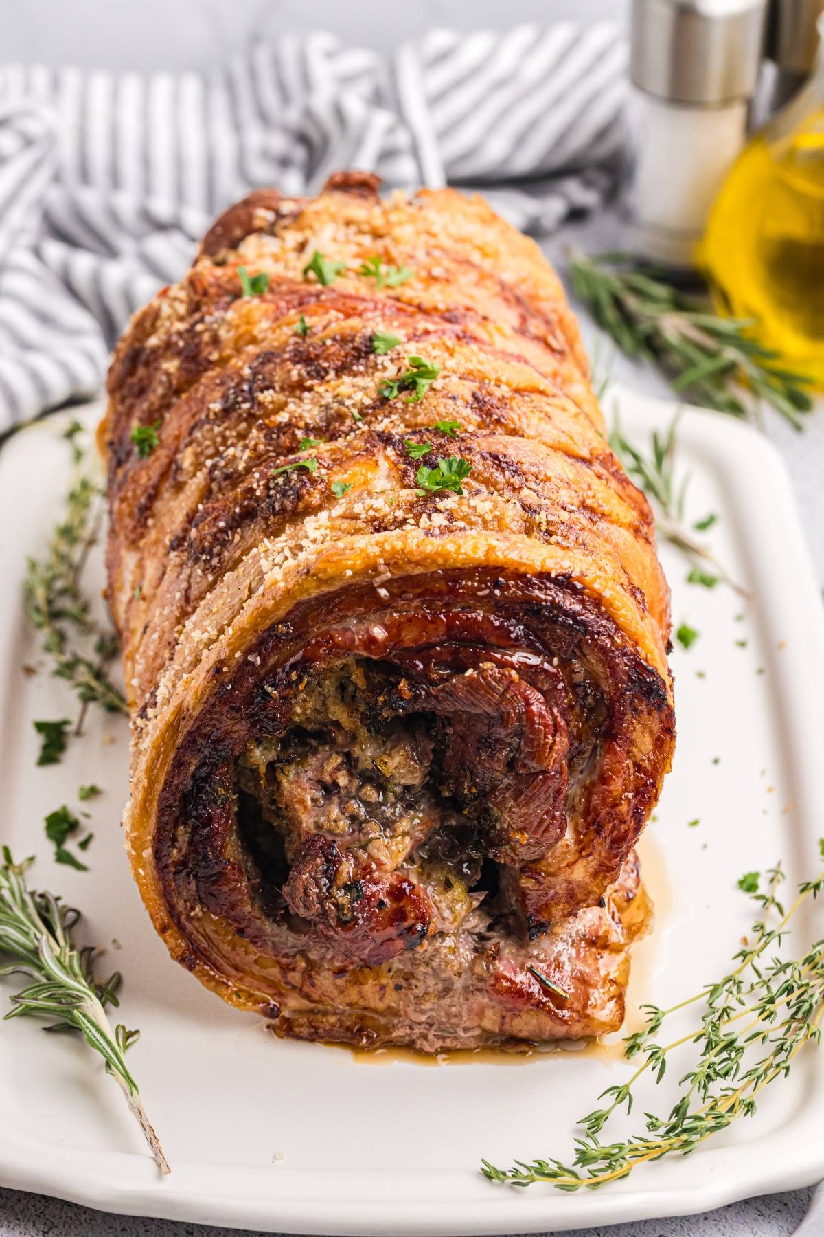 Rolled, roasted pork with crispy skin and herbs on a white platter, garnished with fresh rosemary and thyme.