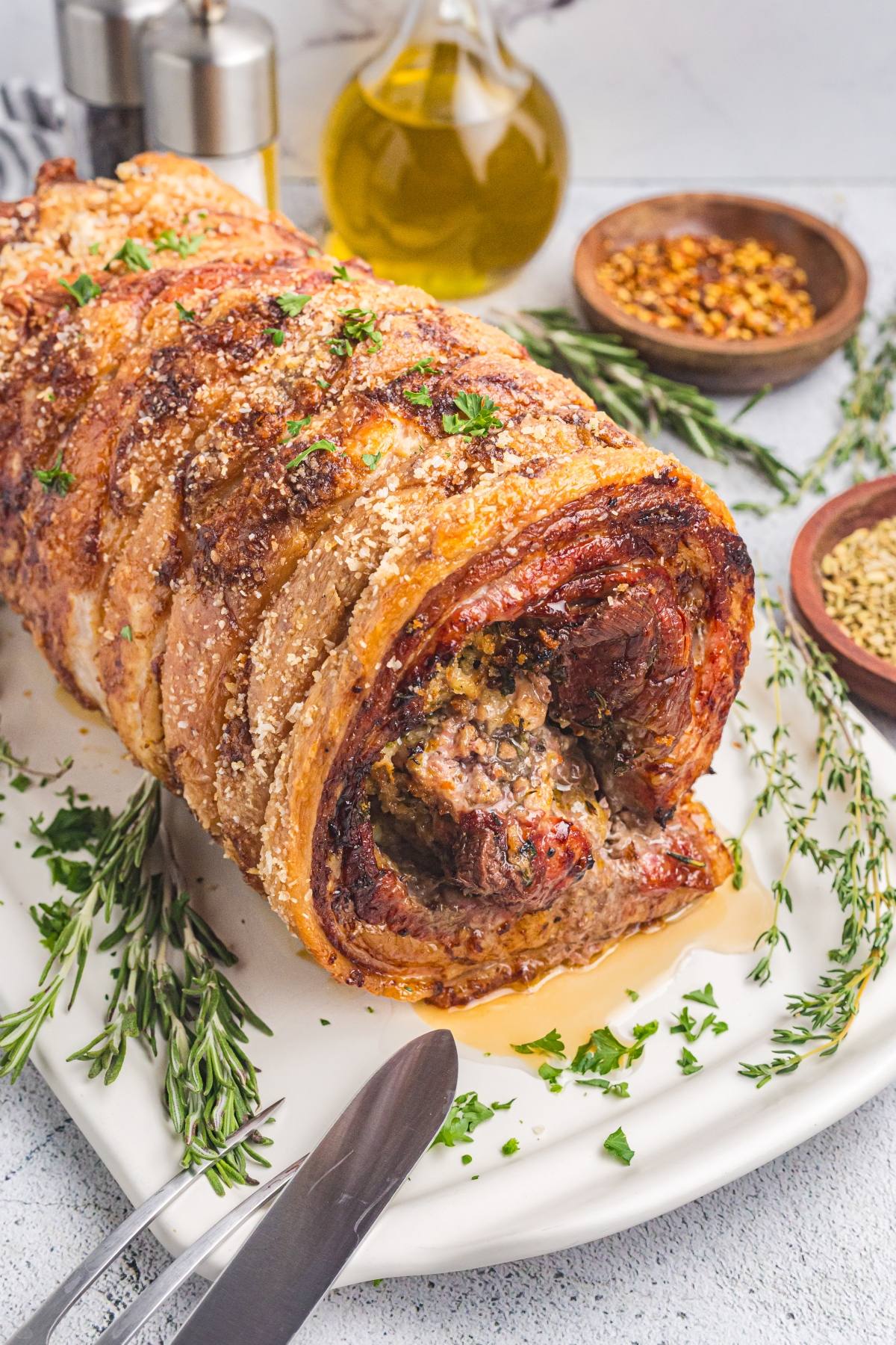 Golden, crispy porchetta roast on a platter, garnished with herbs and surrounded by seasonings and olive oil.