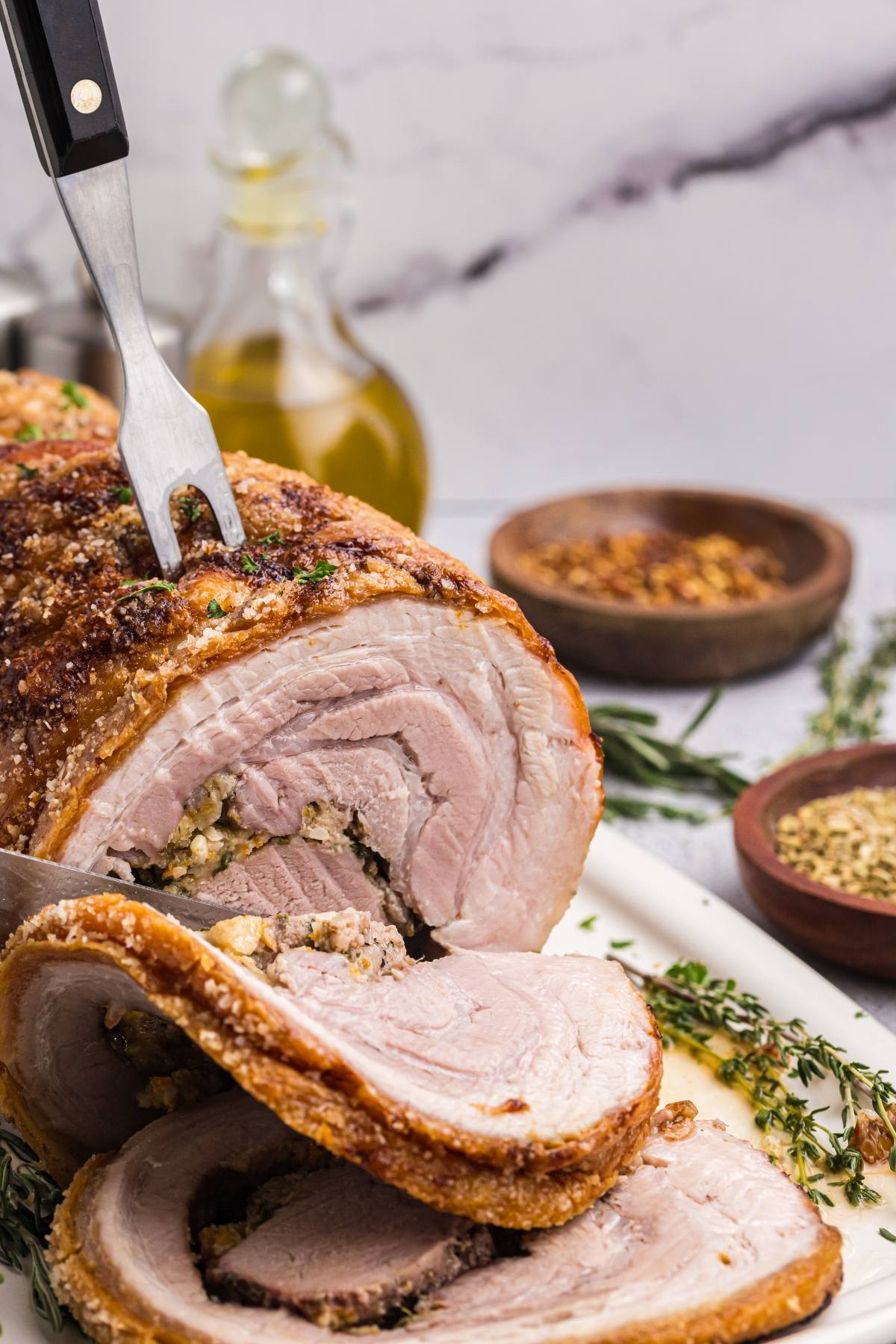 A roasted rolled pork with crispy skin being sliced, garnished with herbs, and seasoned with spices nearby.