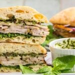 A stacked Porchetta Sandwich with greens on ciabatta bread, served next to a bowl of green sauce and fresh basil.