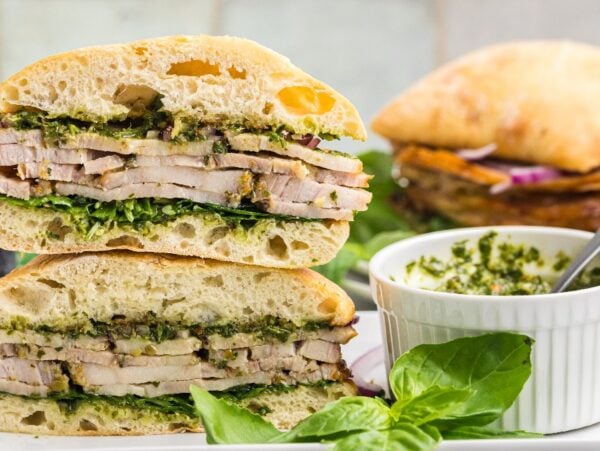 A stacked Porchetta Sandwich with greens on ciabatta bread, served next to a bowl of green sauce and fresh basil.