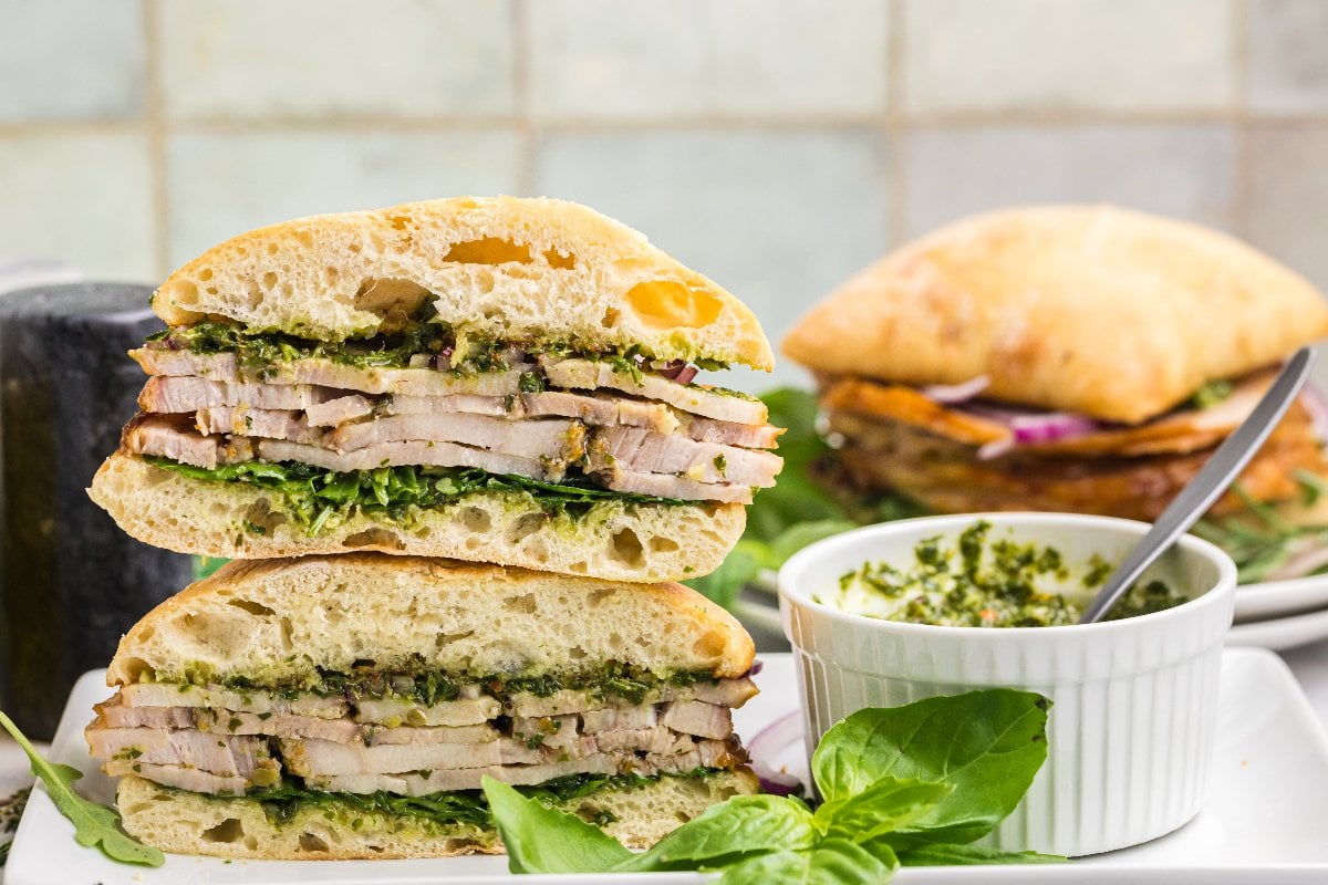 A stacked Porchetta Sandwich with greens on ciabatta, served next to a small bowl of green pesto sauce.