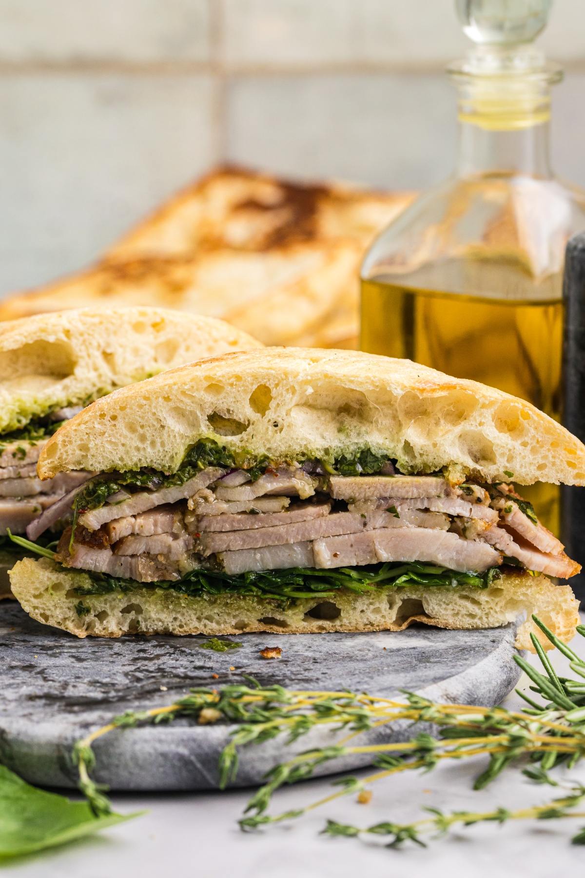 A sandwich with sliced meat, greens, and pesto on ciabatta bread, set on a stone board near an olive oil bottle.