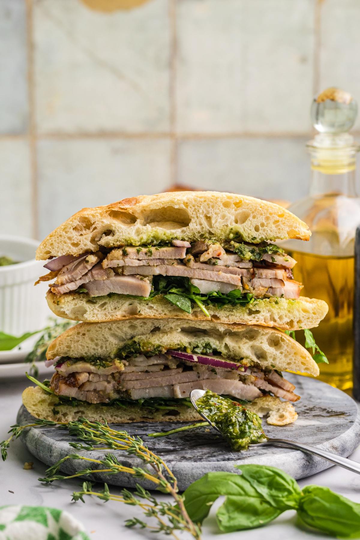 A stacked pork sandwich with greens and pesto on ciabatta bread, set on a marble board with herbs and olive oil.