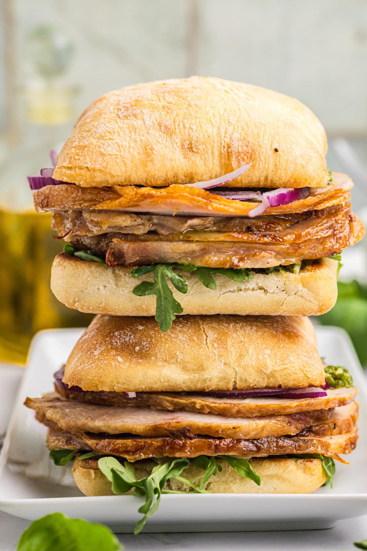 Two stacked ciabatta sandwiches with roast meat, arugula, red onion, and sauce on a white plate.