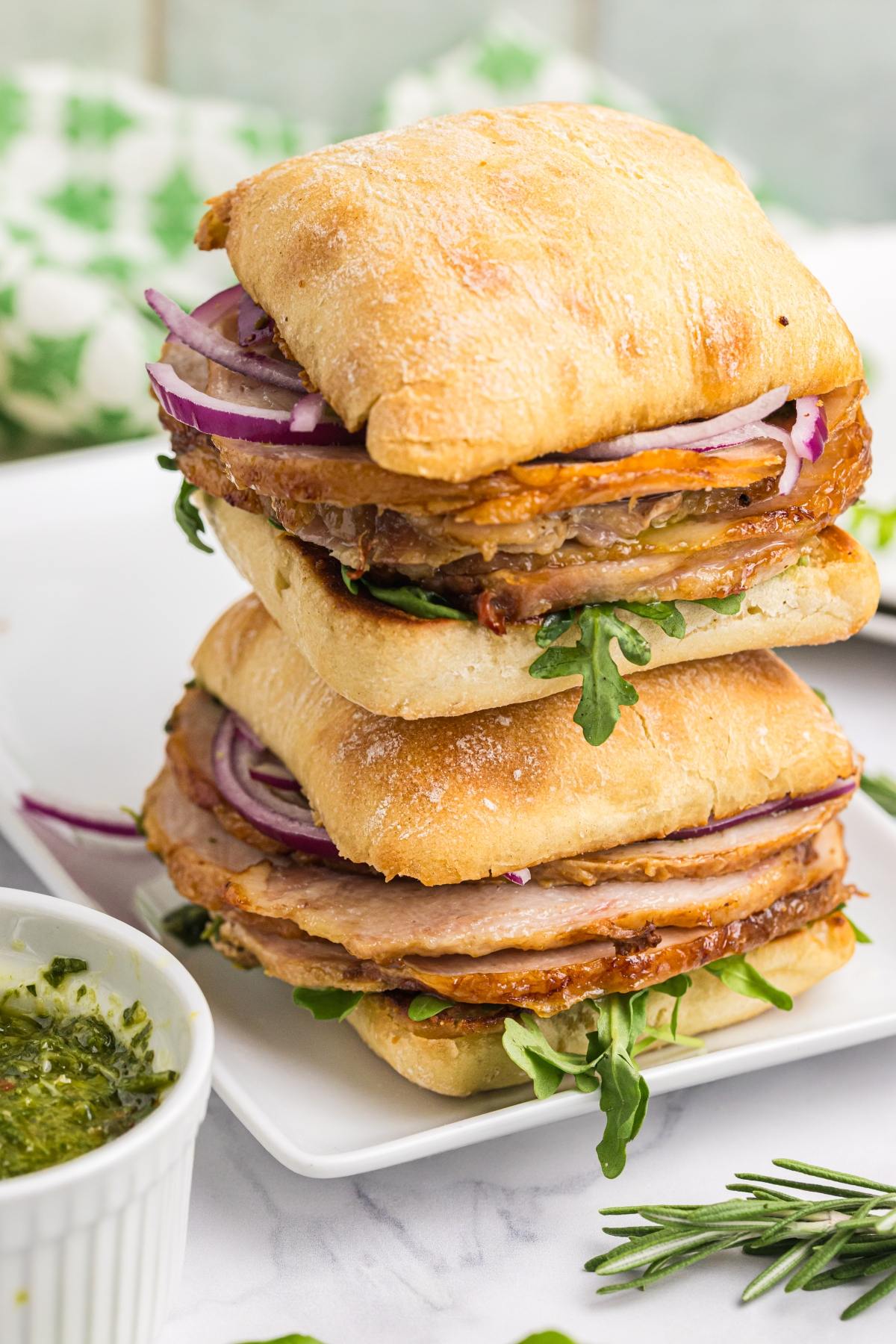 Two stacked ciabatta sandwiches with roast meat, arugula, red onion, and sauce on a white plate.