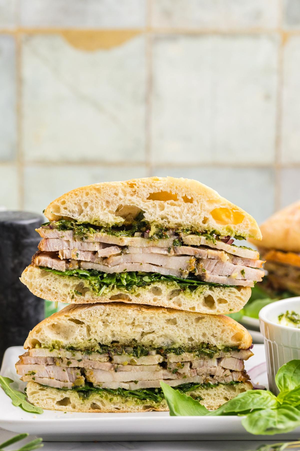 A sandwich cut in half, filled with sliced pork, greens, and sauce, stacked on a plate.