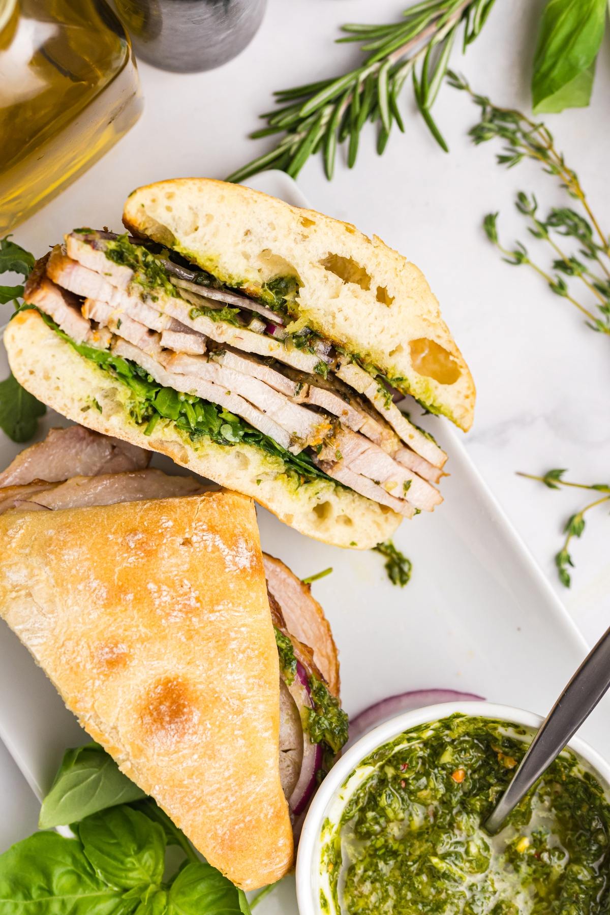 A sliced pork sandwich with green sauce on ciabatta bread, served with herbs and a small bowl of chimichurri.