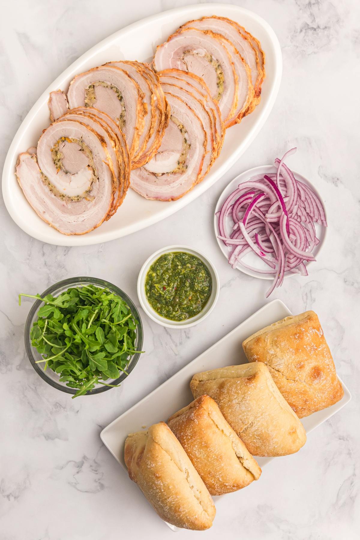 Sliced porchetta, red onion, arugula, chimichurri sauce, and ciabatta rolls arranged on a marble surface.