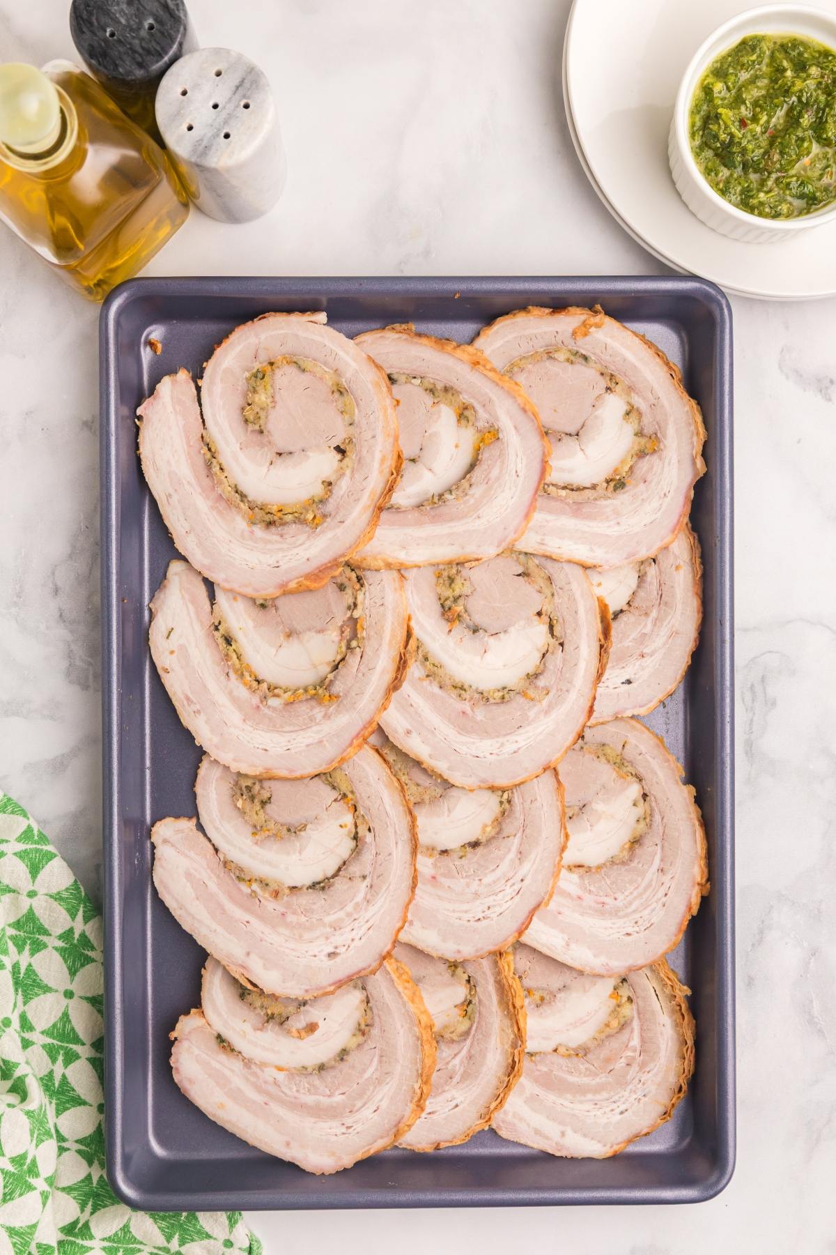 A baking sheet with neatly arranged slices of rolled roasted pork, next to sauce and condiments.