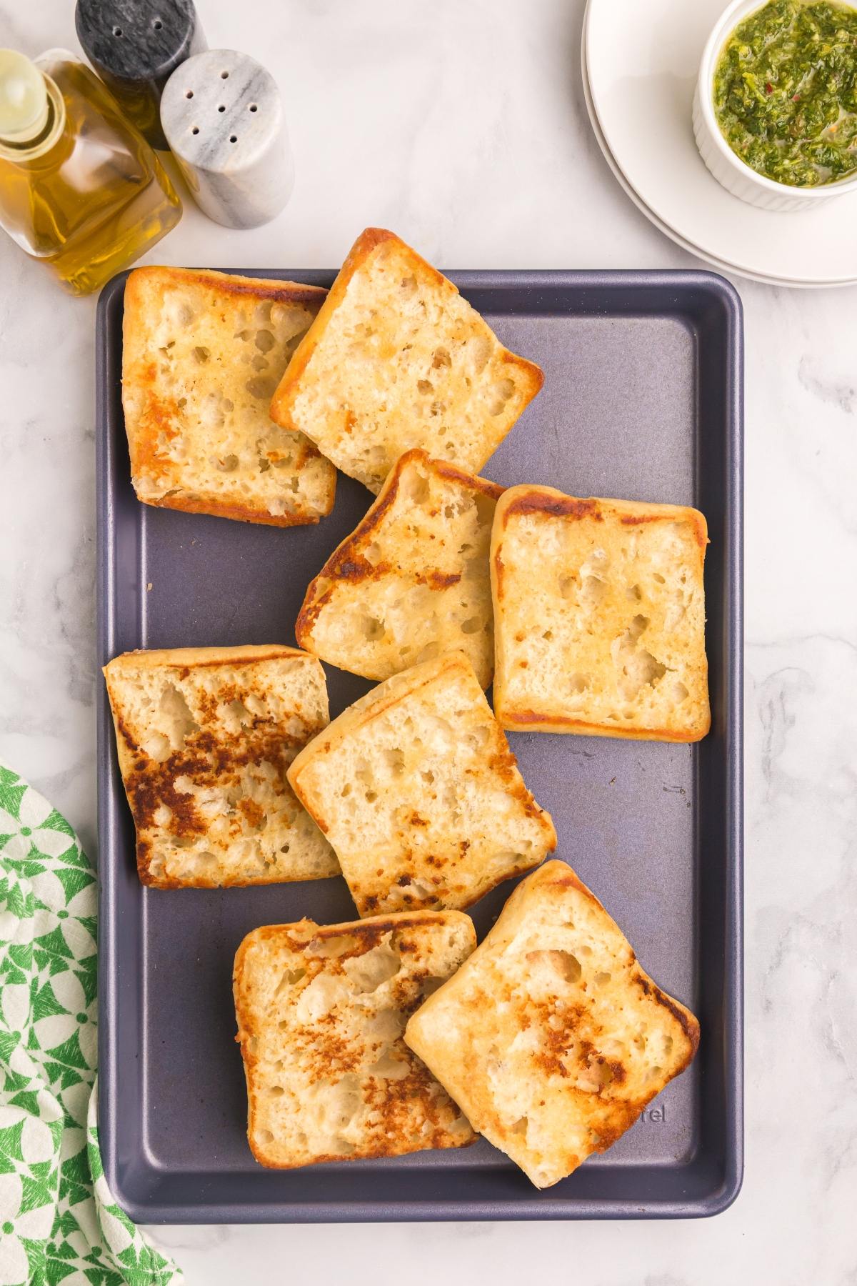 Eight toasted ciabatta bread pieces on a baking sheet, with olive oil and pesto nearby.