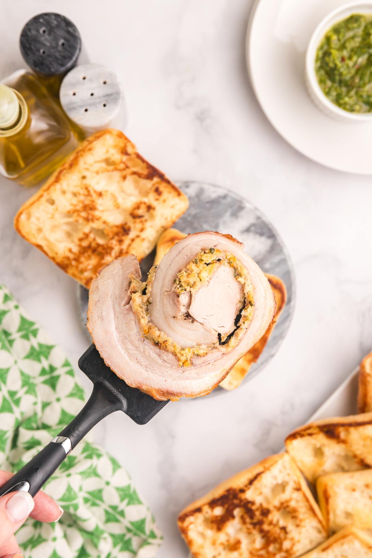 A spatula holds a slice of stuffed pork roast above toasted bread, with herbs and condiments nearby.