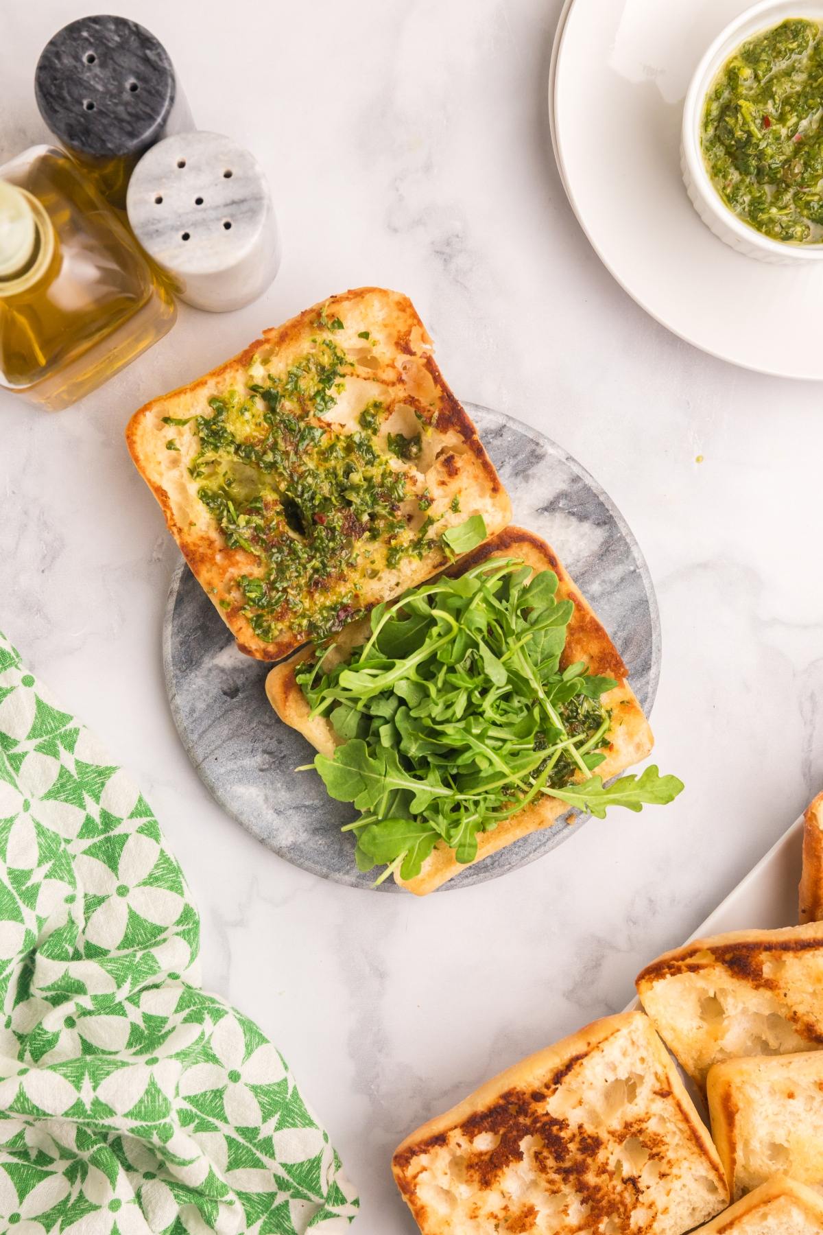 Toasted sandwich bun halves with pesto and arugula on a round board, surrounded by condiments and a napkin.