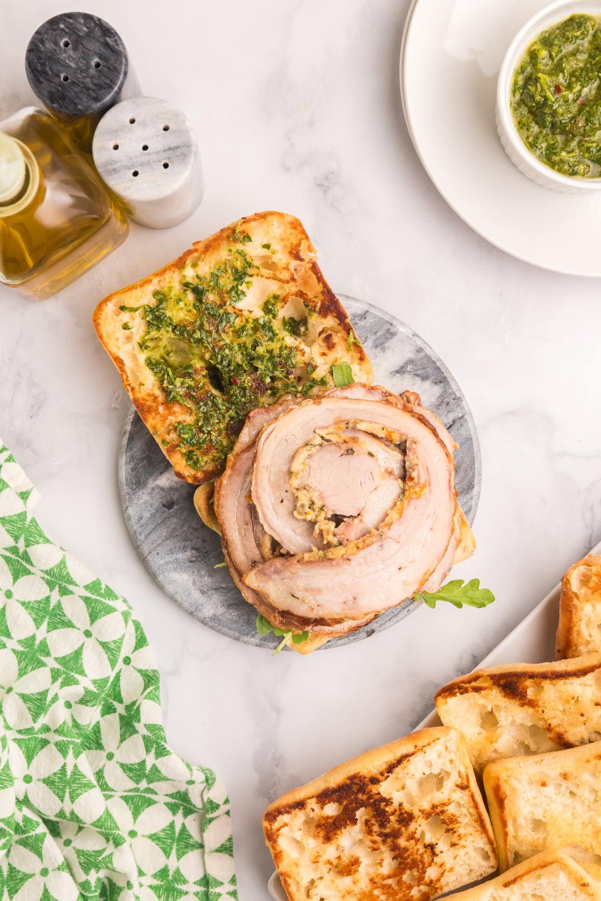 Open porchetta sandwich with green sauce on toasted bread, surrounded by focaccia, olive oil, and a green napkin.