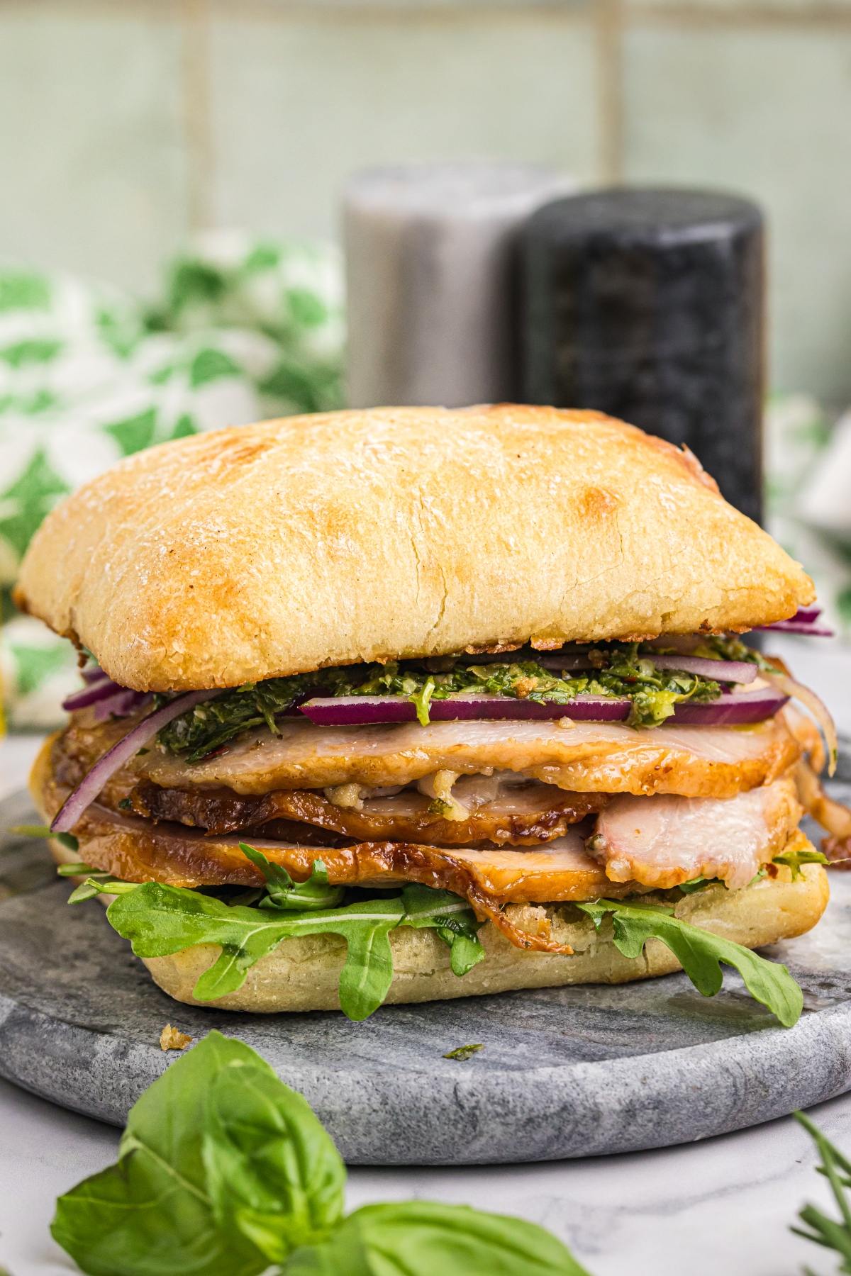 A stacked ciabatta sandwich with grilled chicken, red onions, arugula, and pesto on a stone board.