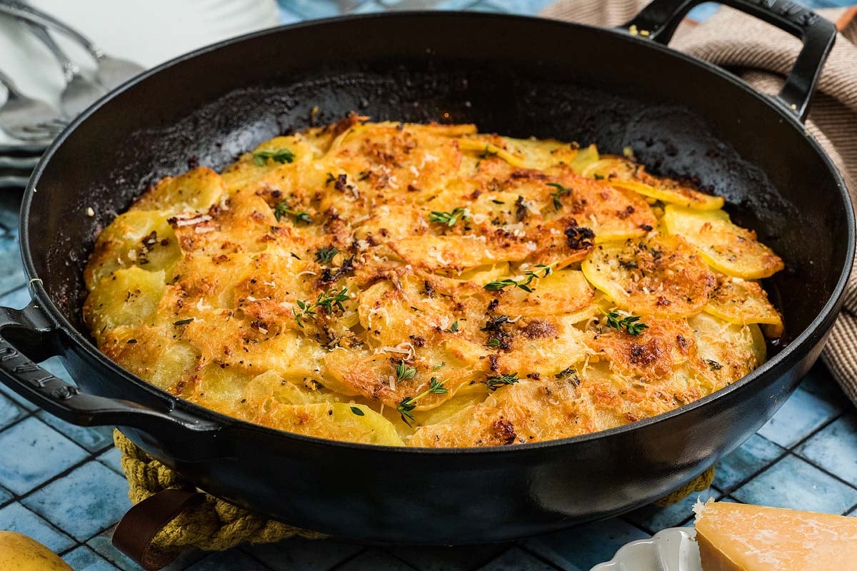 A cast iron skillet filled with golden, baked Potato Galette, garnished with fresh herbs.