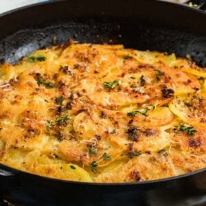 Golden, baked potato galette topped with herbs in a black cast iron pan on a blue tiled surface.