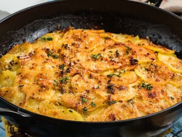Golden, baked potato galette topped with herbs in a black cast iron pan on a blue tiled surface.