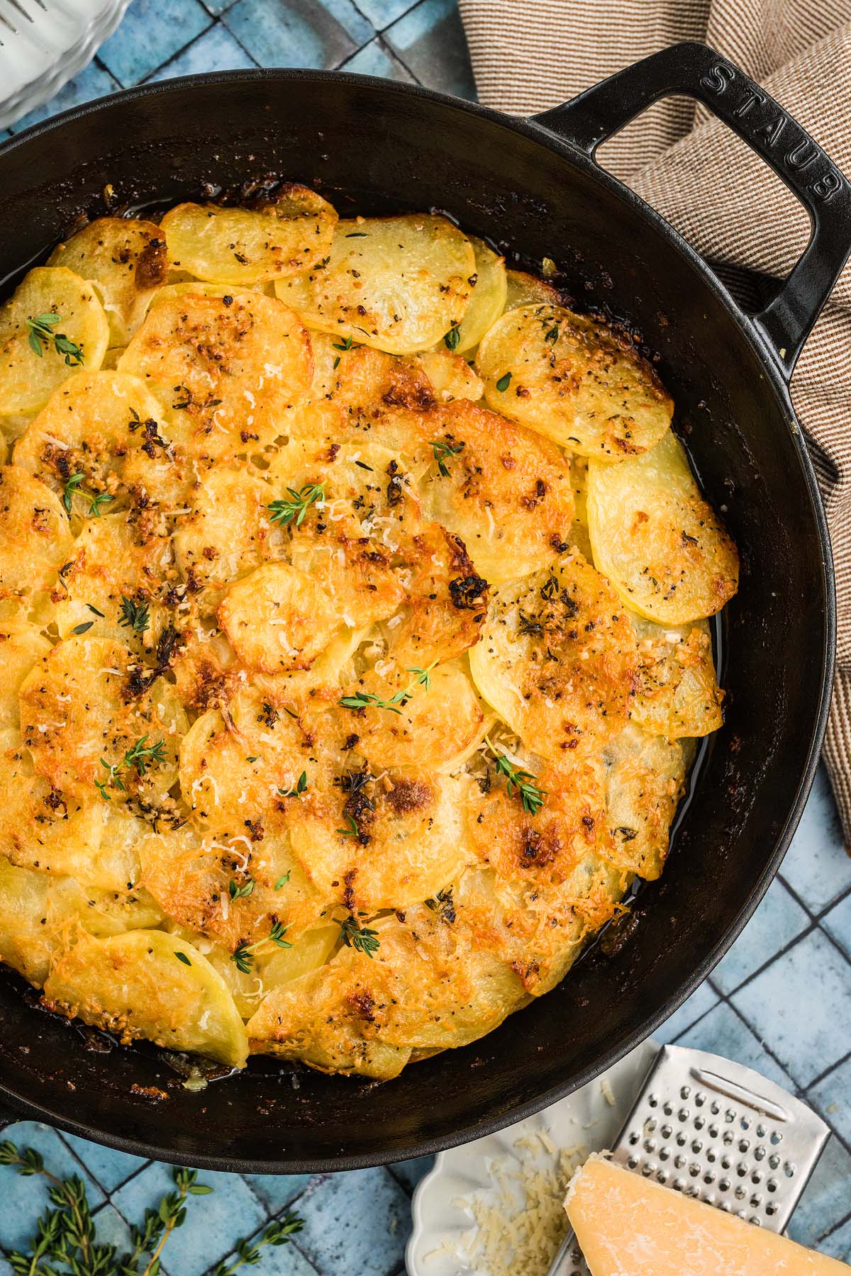 Golden, crispy Potato Galette topped with fresh herbs in a black skillet on a blue tiled surface.
