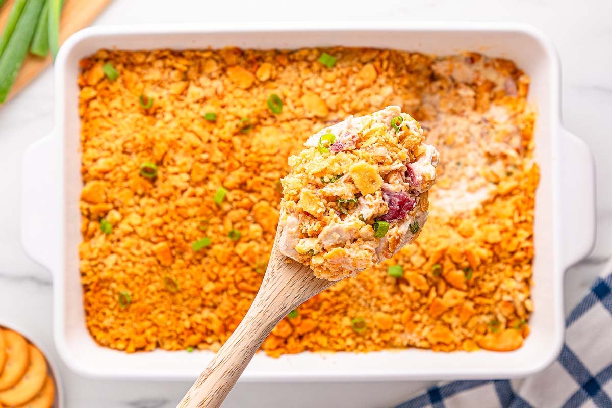 A wooden spoon holds a serving of golden, cheesy Ritz Cracker Chicken Casserole with a crunchy topping above a baking dish.